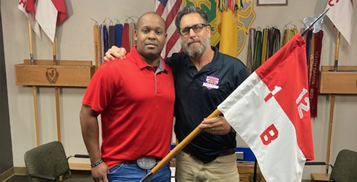 Veterans holding a unit flag in front of military insignia, honoring their service and the stories behind current projects.