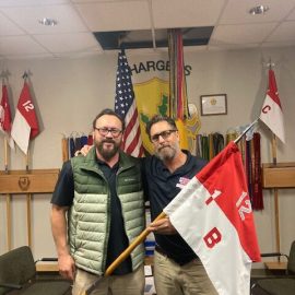 Veterans holding a unit flag in front of military insignia, honoring their service and the stories behind current projects.